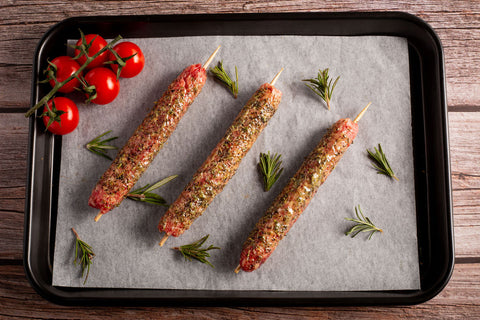 Three lamb skewers of seasoned meat on a baking tray with fresh tomatoes and rosemary.