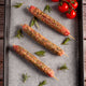 Three lamb skewers of seasoned meat on a baking tray with fresh tomatoes and rosemary.