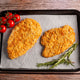 Two breaded chicken fillets on a baking tray with tomatoes and rosemary on a wooden surface