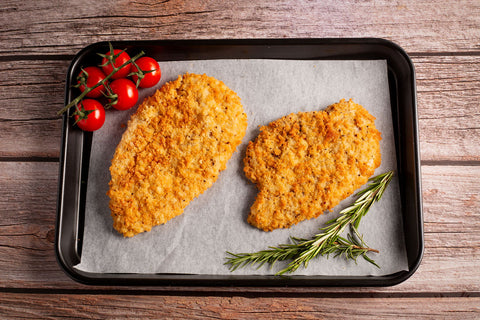 Two breaded chicken fillets on a baking tray with tomatoes and rosemary on a wooden surface